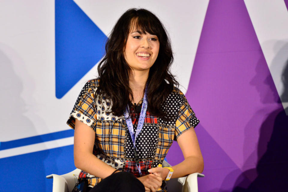 Jaiden smiles while seated in front of a geometric backdrop. She wears a multi-patterned shirt and a lanyard