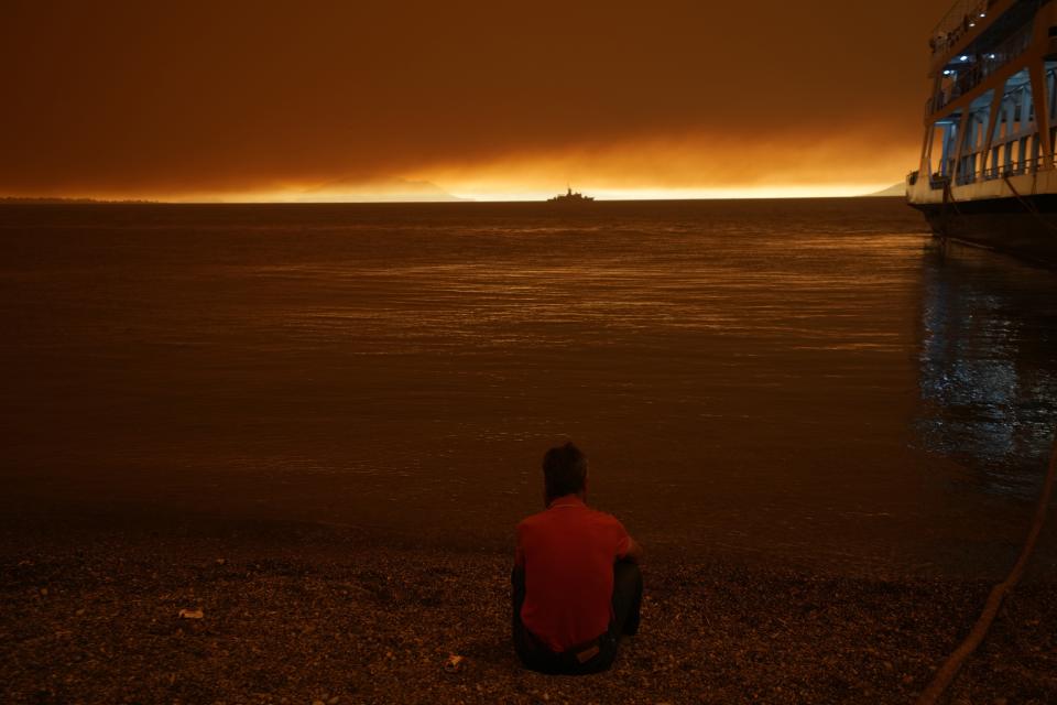 A man sits next to a ferry which used to evacuate from Pefki village on Evia island, about 189 kilometers (118 miles) north of Athens, Greece, Sunday, Aug. 8, 2021. Pillars of billowing smoke and ash are blocking out the sun above Greece's second-largest island as a days-old wildfire devours pristine forests and triggers more evacuation alerts. (AP Photo/Petros Karadjias)