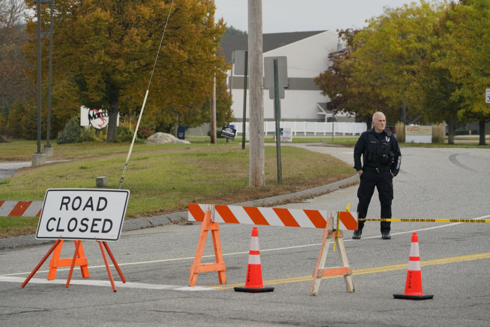 A police officer stands at a road closure near a bowling alley, seen in background, Thursday, Oct. 26, 2023, in Lewiston, Maine. The site is one of Wednesday’s two mass shootings in the city. Residents have been ordered to shelter in place as police continue to search for the suspect. (AP Photo/Robert F. Bukaty)