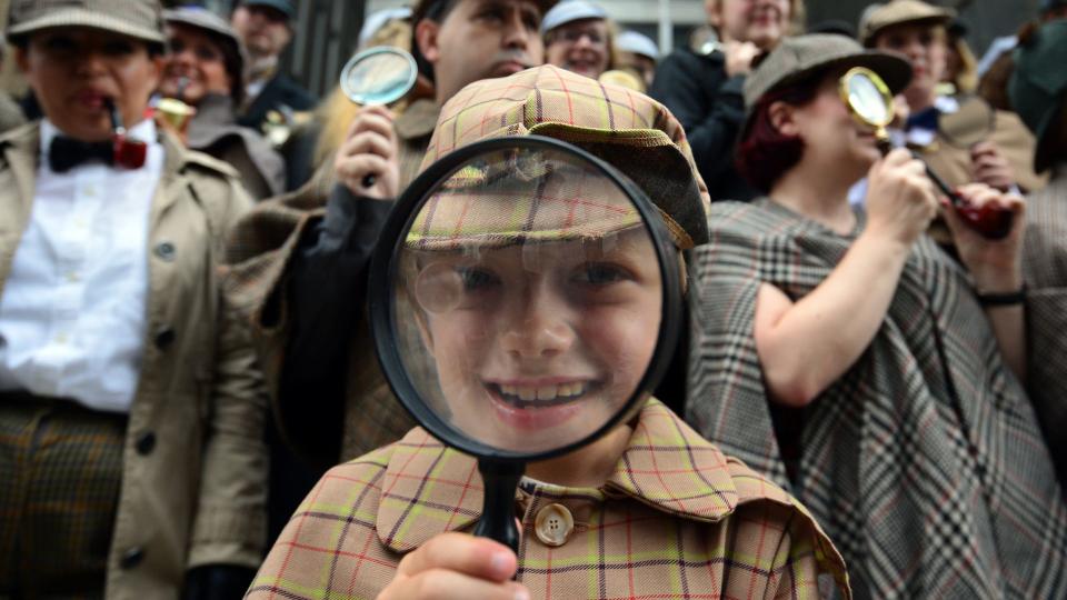 close up on a boy dressed as sherlock holmes and looking through a magnifying glass surrounded by other children and adults also dressed as sherlock holmes in central London in Summer 2014