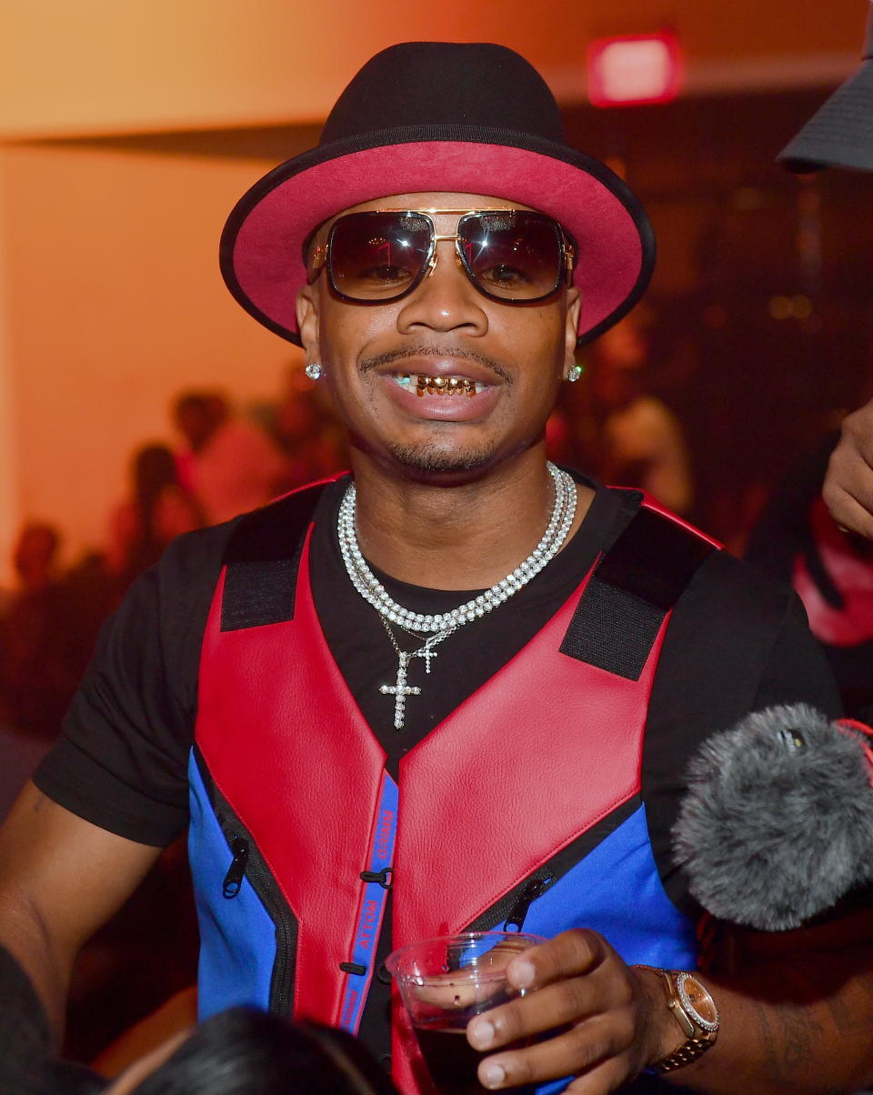 Pryce, wearing sunglasses, a red and black hat, a red and blue vest and a diamond necklace, is seen holding a drink at the event.