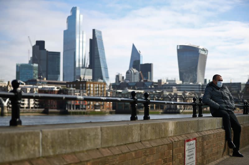 A man wearing a protective mask sits along the Southbank in front of the City of London financial district during rush hour, as the number of Coronavirus cases grow around the world, in London