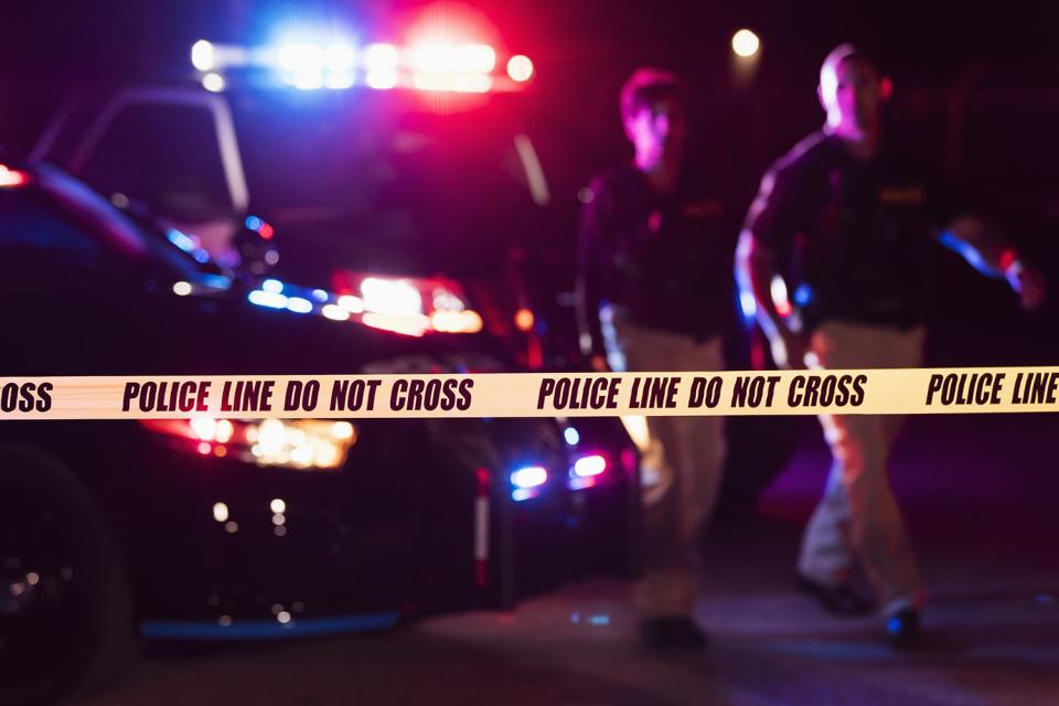 <p>Getty</p> Two police officers standing in front of patrol cars, behind police tape.