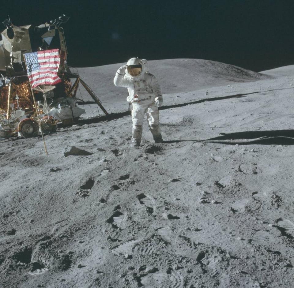 an astronaut in a bulky white spacesuit plants an american flag on the dusty grey surface of the moon