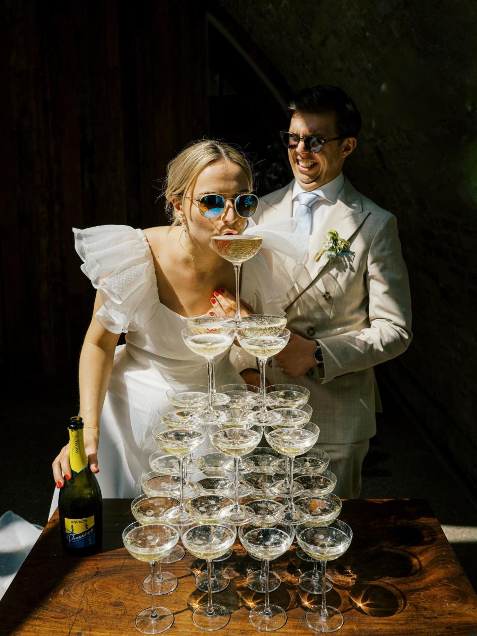 A bride sips champagne from the top glass of a champagne tower.