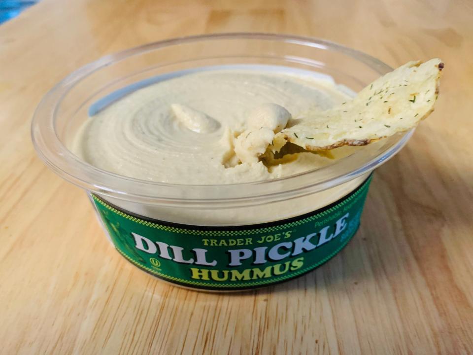 A chip in an open container of Trader Joe's dill-pickle hummus on a light wood table