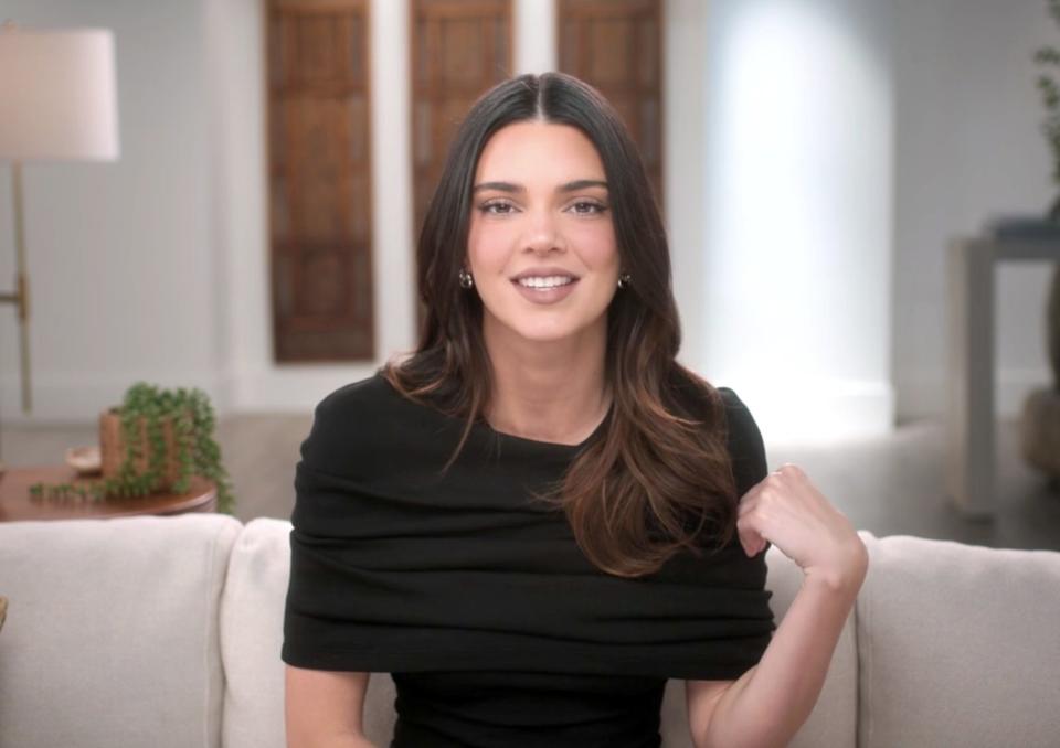 Kendall Jenner sitting on a couch, smiling and wearing a stylish top