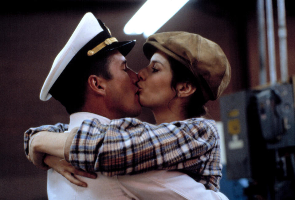 Richard Gere and Deborah Winger share a kiss, with Gere in a naval uniform and Winger wearing a plaid shirt and a cap