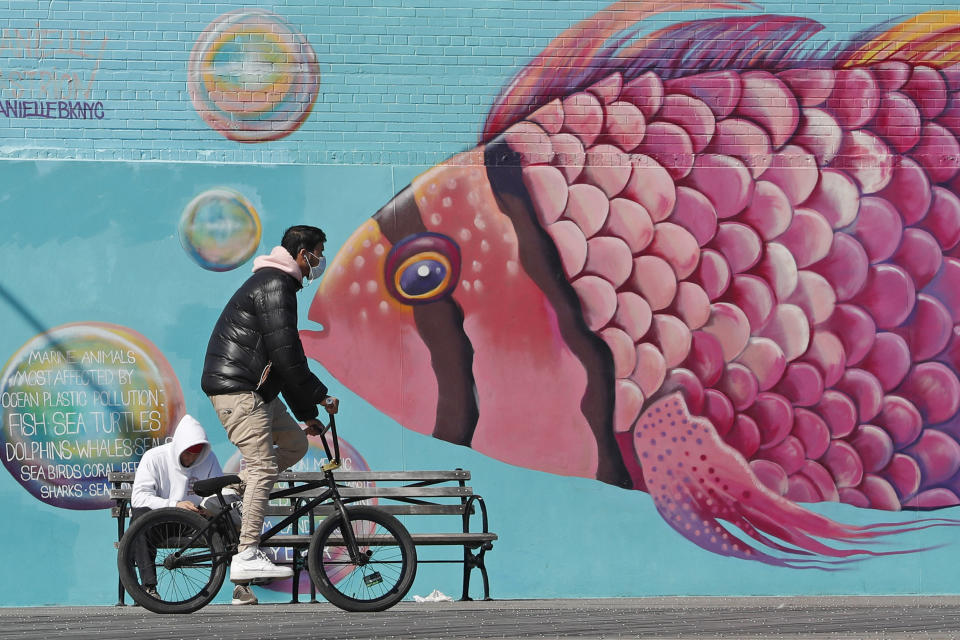 FILE - In this April 2, 2020, file photo, a face-mask wearing cyclist pedals past a mural of a tropical fish on the Coney Island boardwalk in the Brooklyn borough of New York. From Cape Cod to California, festivals are being canceled, businesses in tourist havens are looking at empty reservation books, and people who have been cooped up through a dismal spring are worrying summer will bring just more of the same. (AP Photo/Kathy Willens, File)