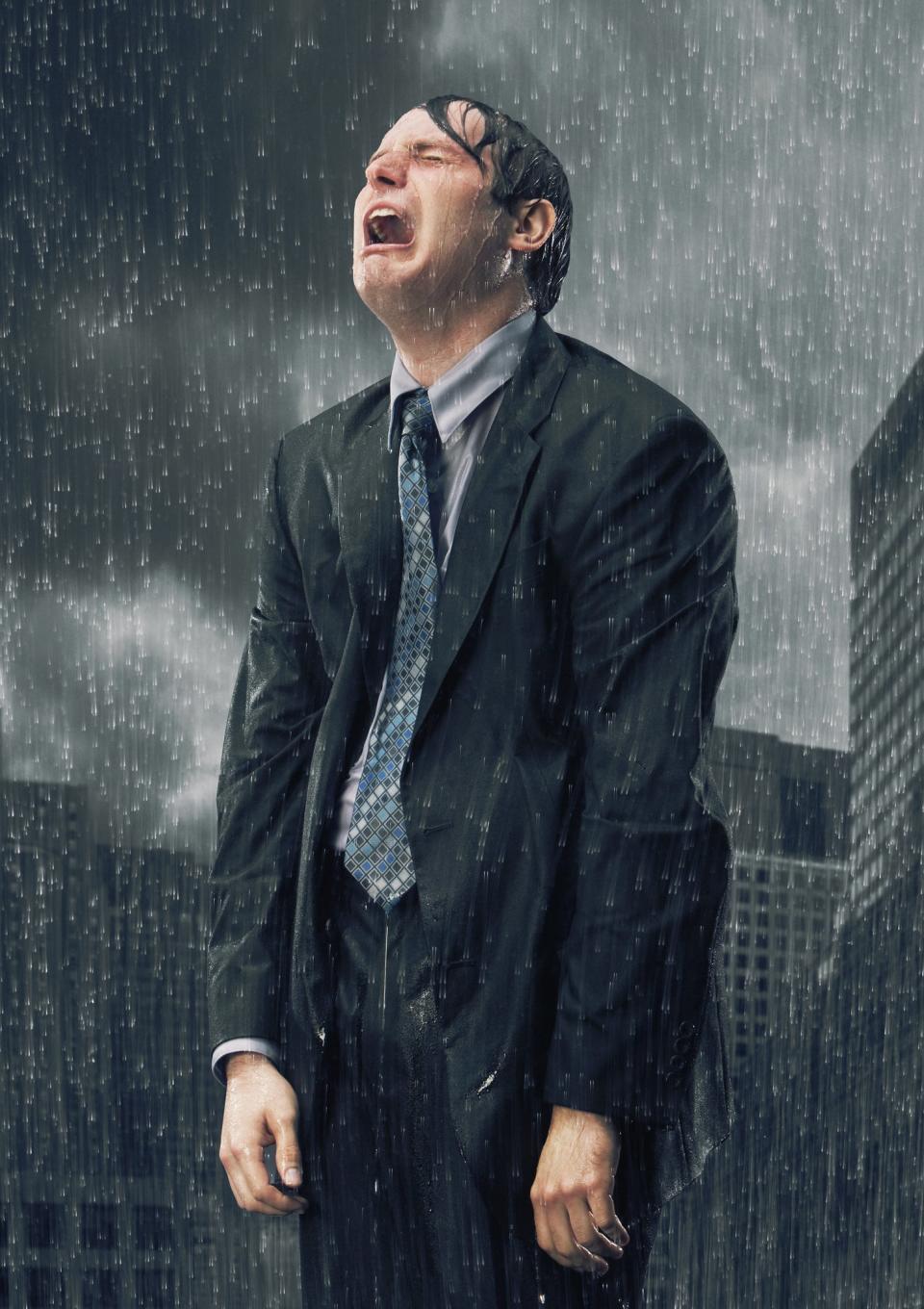 Man in suit stands in rain with eyes closed and mouth open, as if catching raindrops. City buildings in background