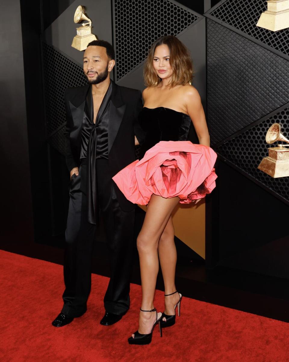 John Legend in a black suit with satin details. Chrissy Teigen in a short black dress with a huge pink flower at her hip