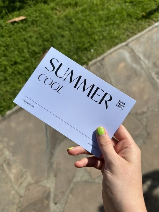 A hand with green nail polish holds a card reading "SUMMER COOL, OMYO COLOR STUDIO" on an outdoor stone pavement next to grass