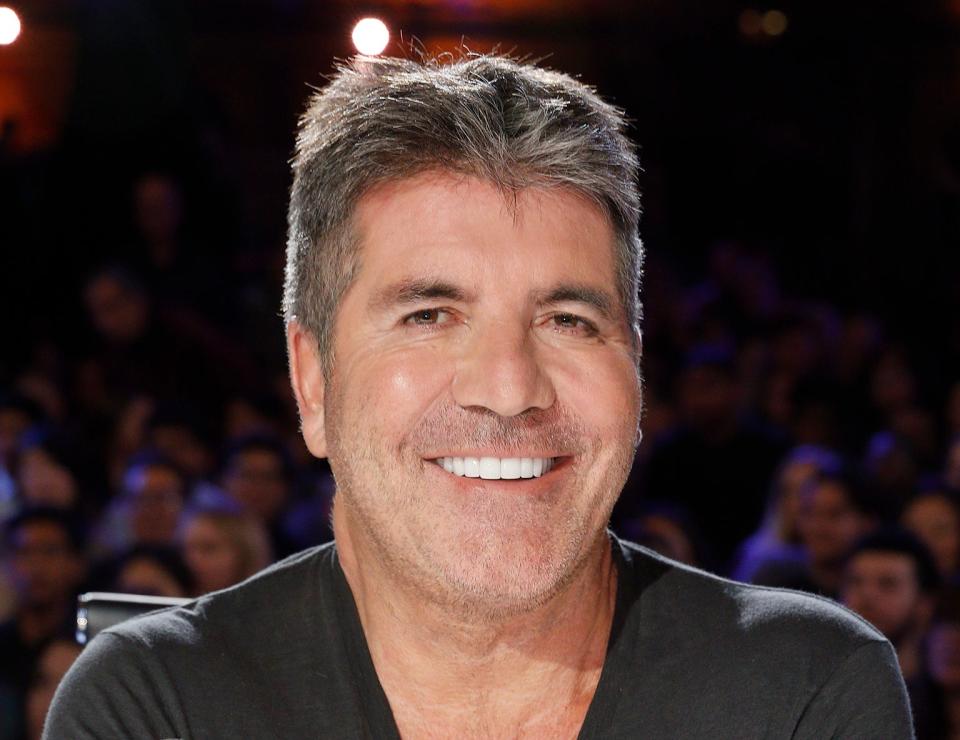 Simon Cowell smiling at a judging table with a microphone and buzzer