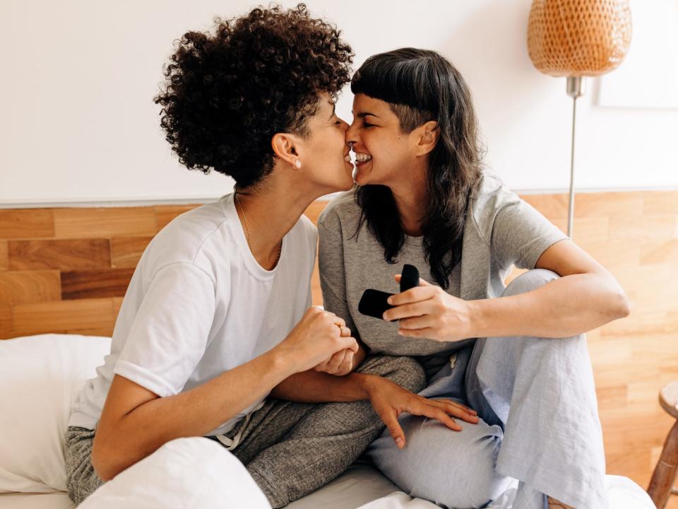 Two women kiss in bed as one holds an engagement ring box.