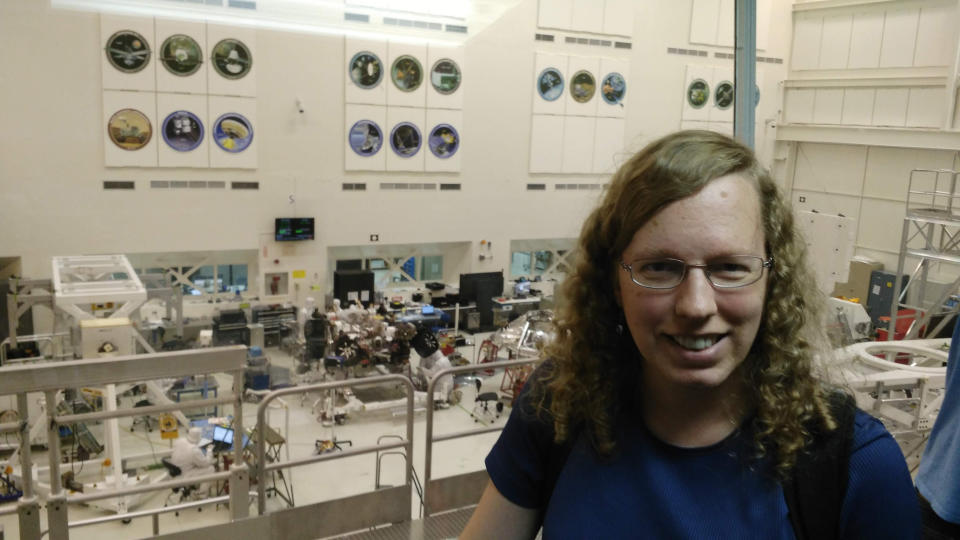 a woman in glasses stands overlooking technicians working on a space rover in a white room.