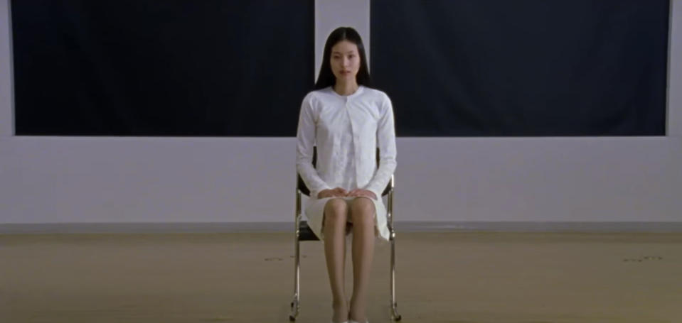 A woman, dressed in a simple, long-sleeved white dress, sits alone on a chair in a minimalist setting; her hands are placed on her knees