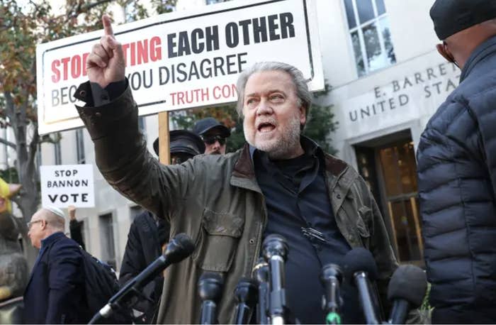 Steve Bannon standing outside a courthouse pointing up at the sky in front of a bunch of reporter mics