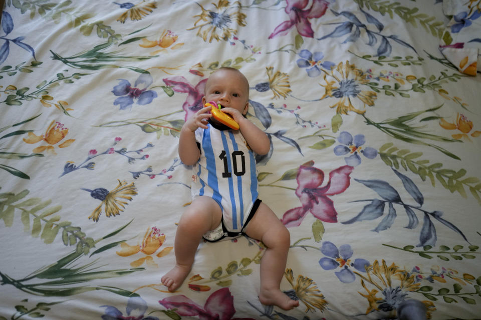 Wearing an Argentina inspired national soccer team onesie, Lev Andres mouths a baby rattle while lying on a his parents’ bed, in Mendoza, Argentina, Tuesday, Feb. 14, 2023. Over the past year, Argentine immigration authorities have noticed flights packed with dozens of pregnant Russians. But whereas Lev’s mother said her and her husband intend to build a life in Argentina, local officials believe many of the other recent Russian visitors are singularly focused on receiving one of Argentina’s passports. (AP Photo/Natacha Pisarenko)