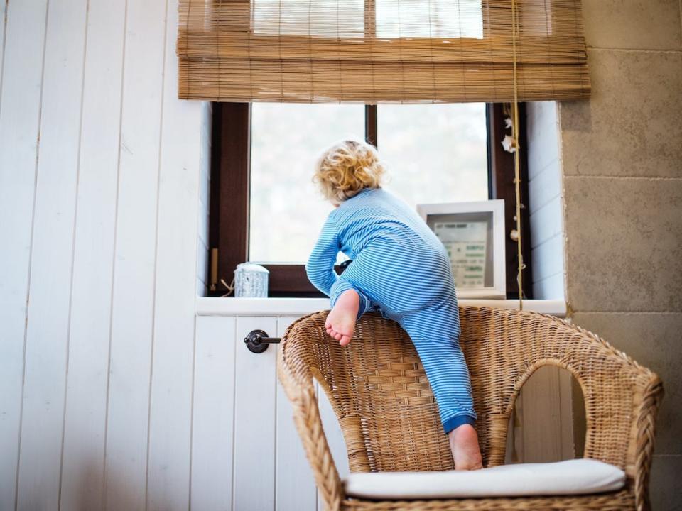 A baby crawls onto a chair and peers out a window.