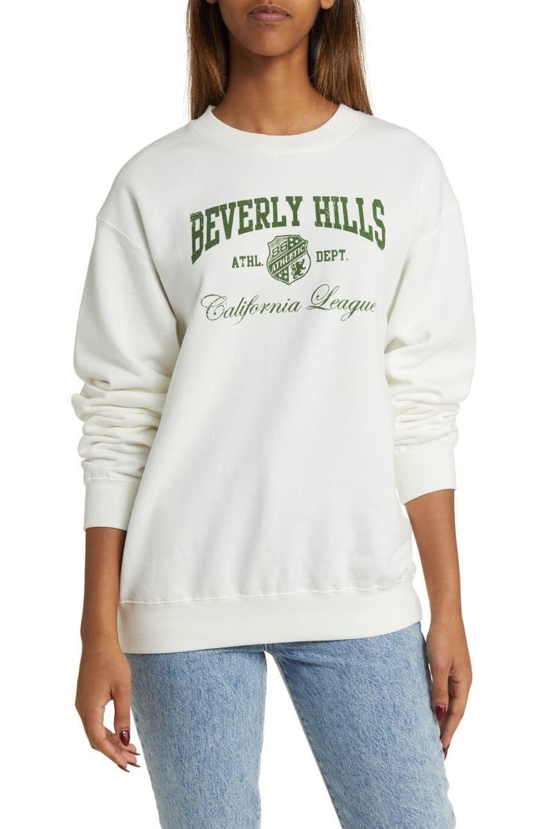 model wearing white sweatshirt with green letters that say "Beverly Hills" on front
