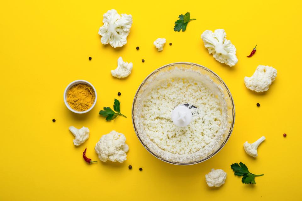 Blender bowl with freshly prepared cauliflower rice and spices on yellow background.