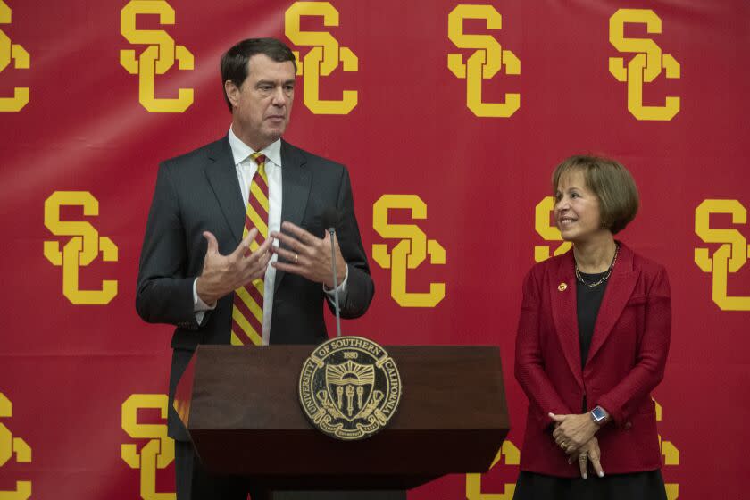 Mike Bohn stands at podium and speaks to a crowd while Carol Folt looks at him and smiles