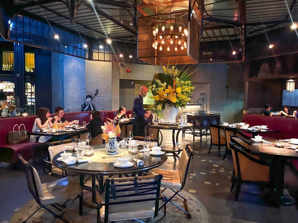 Dark dining area with black tables, black chairs with white cushions, and red chairs. A table with a yellow flower arrangement sits at the center of the room as a table filled with people eats and a waiter walks next to the flowers.