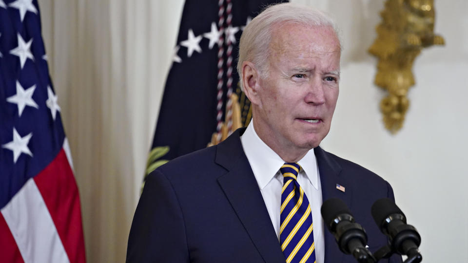 Joe Biden speaking into a microphone with an American flag behind him.