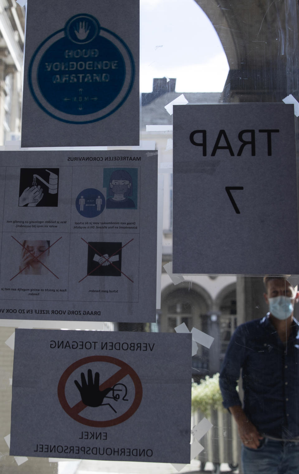 A teacher wearing a protective face mask looks through a window with notices about coronavirus, COVID-19 at the Atheneum high school in Antwerp, Belgium, Monday, Aug. 31, 2020. Students return to school in Belgium on Tuesday, Sept. 1, with measures in place regarding social distancing and the wearing of masks to protect against the spread of coronavirus, COVID-19. (AP Photo/Virginia Mayo)