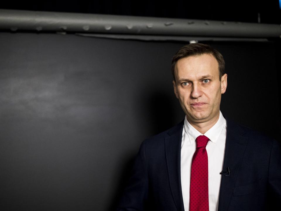 Alexei Navalny wears a suit and a red tie in front of a black wall.
