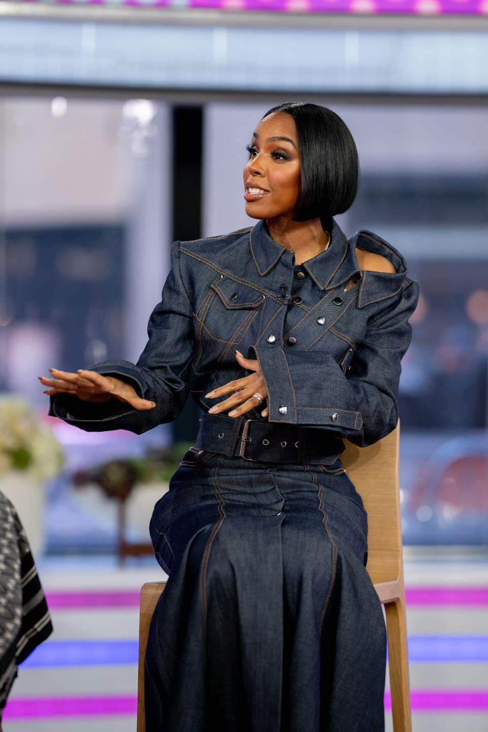 Kelly in a stylized denim jacket and skirt speaks while sitting and gesticulating with her hand