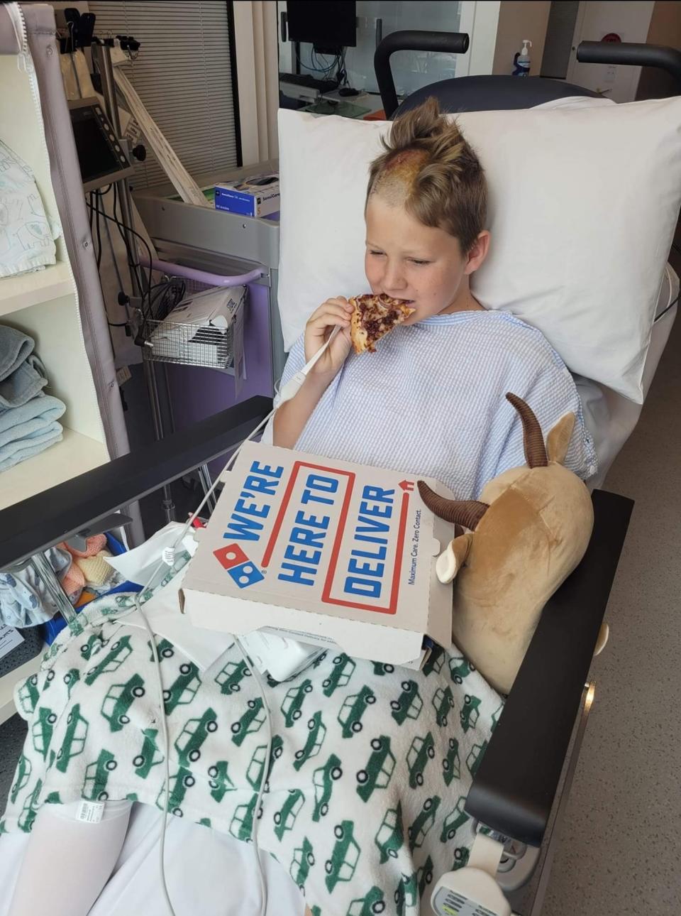 Declean in hospital bed with partly shaved head eating pizza.
