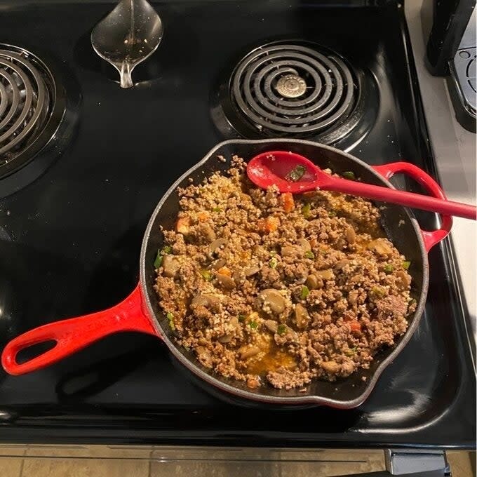 A skillet with ground meat and vegetables on a stove, next to a red spoon