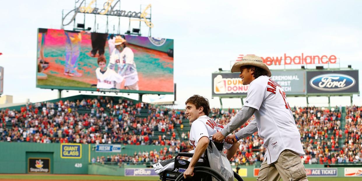 jeff bauman and carlos arredondo, wearing boston red sox jerseys, on the grass of a baseball field, with spectators in seats in the background and their image on a large television screen