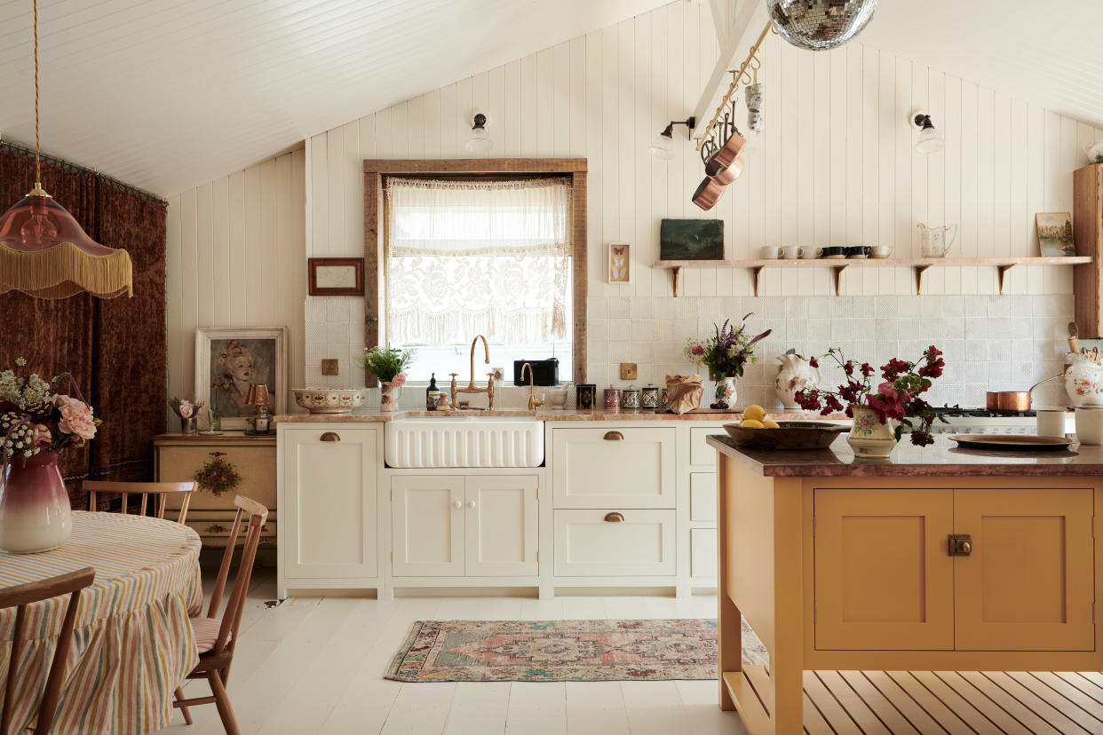  grandmillennial style cottage kitchen with shiplap walls and ceiling, mustard kitchen island, round table and chairs, open shelving, vintage rug, white floorboards, fringed pendant, net curtain at window, copper pans  