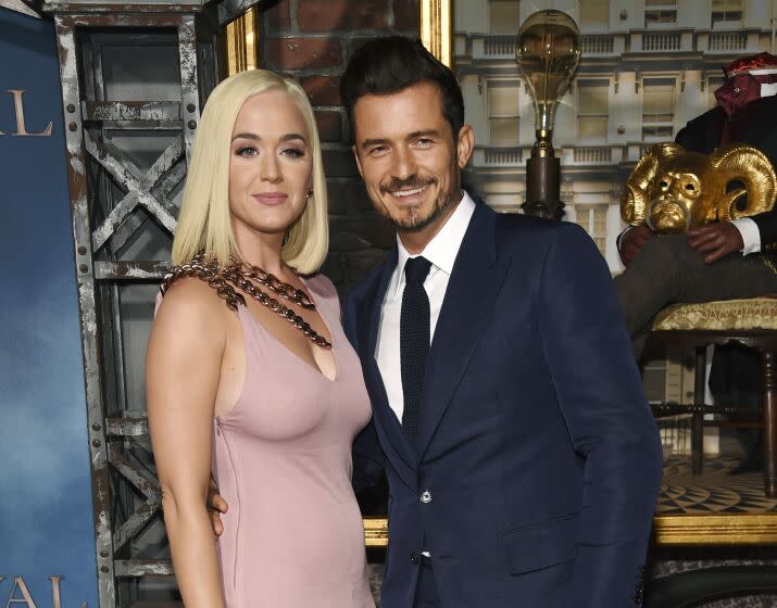 Katy Perry with blonde hair in a pastel pink dress posing next to Orlando Bloom with dark hair and blue suit