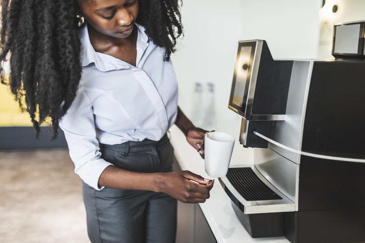 Young adult black woman taking a break at  office and making tea using coffee machine for hot water. Coffee break during hard working day.