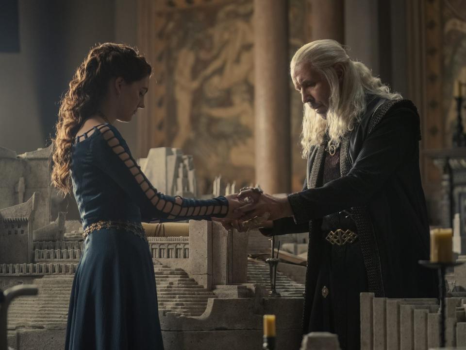 A teen girl in a blue dress holds a stone dragon out to an older man with long silver hair.