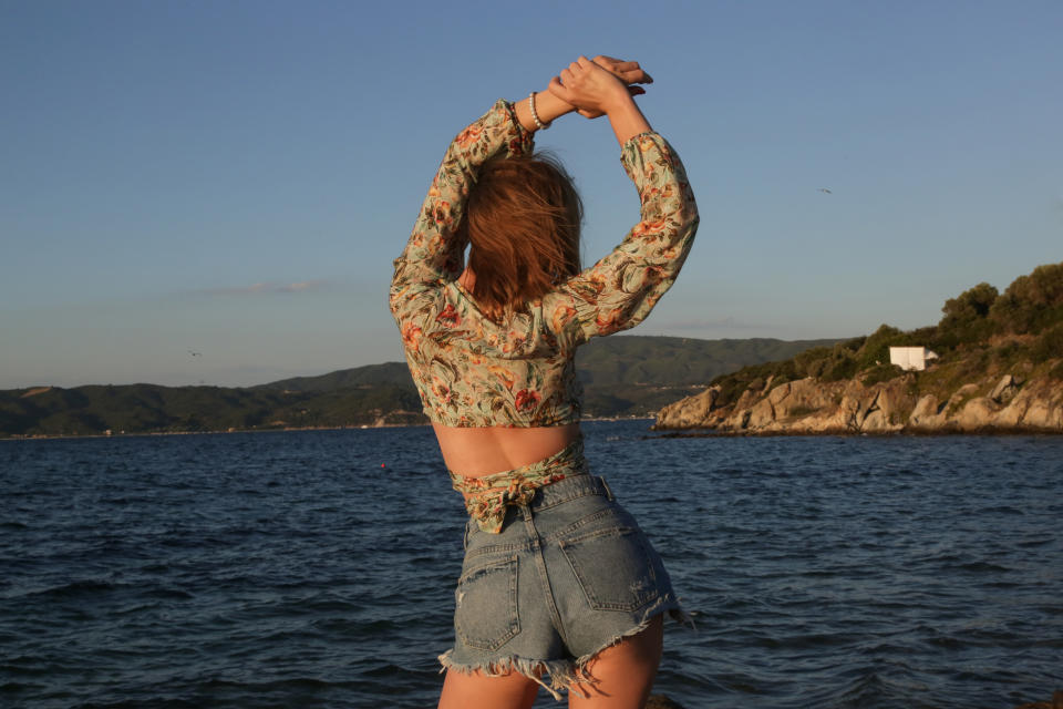 Outdoor portrait of young fashionable woman in crop top and denim shorts with blue sea and clear sky behind.