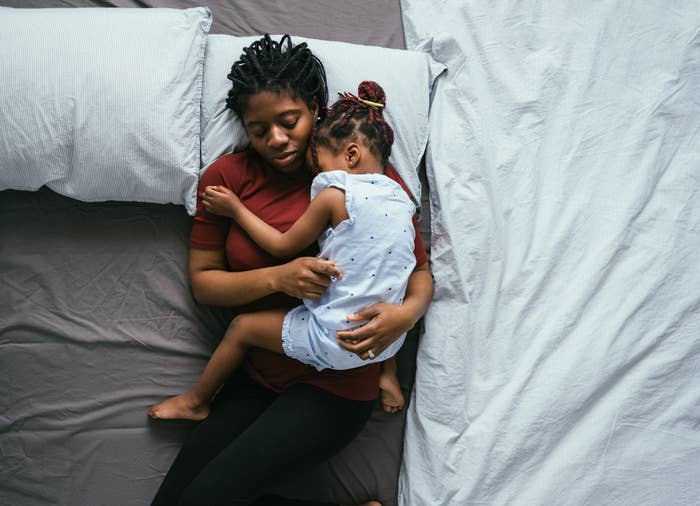 An adult and a child cuddle while sleeping on a bed. The adult is wearing a short-sleeve shirt and leggings. The child is dressed in a short-sleeve dress