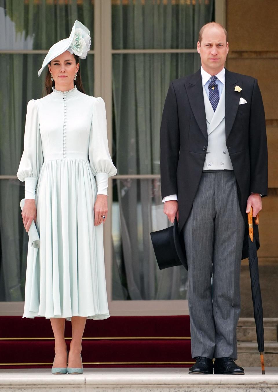 Duchess Kate and Prince William stand at attention for the national anthem before plunging into the crowd at the latest royal Garden Party at Buckingham Palace on May 25, 2022. Kate wore a pale blue-green dress with a high collar, buttons down the bodice and full skirt and sleeves.