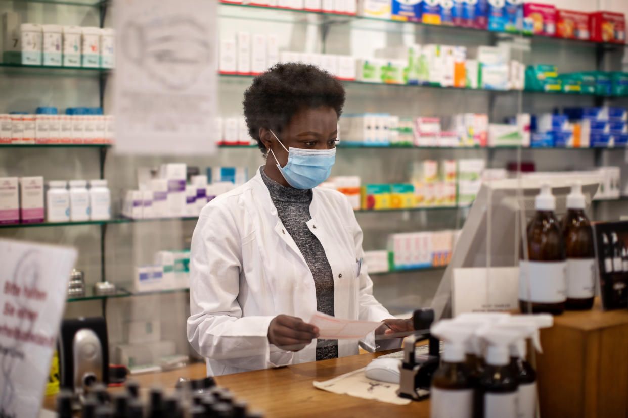 Woman pharmacist wearing surgical face mask checking out customer's medication prescription. African female chemist standing behind the counter  with a screen partition and using computer in medical store.