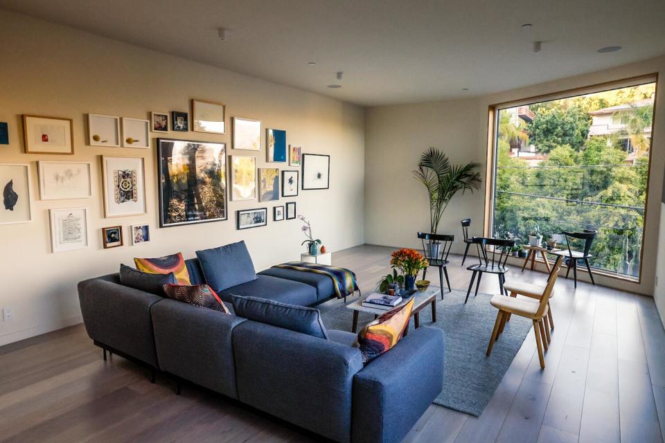 A blue couch sits in the living room near a wall covered in frames and a large window with a view of a green hillside.