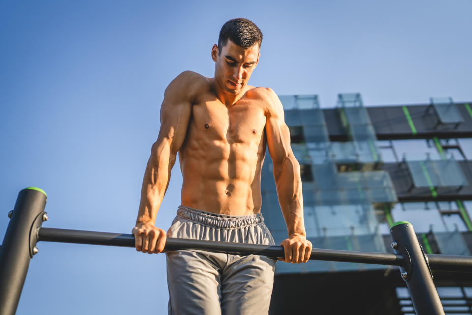 Man performing a muscle-up on an outdoor gym bar, showcasing his muscular physique. He is shirtless, wearing athletic shorts, in front of a modern building