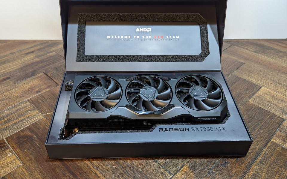 A picture of a black AMD Radeon RX 7900 XTX with its packaging on a brown wooden table.