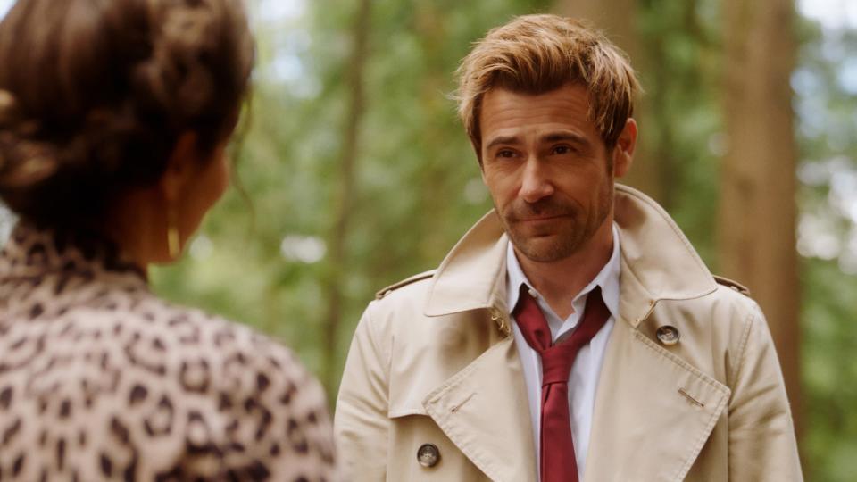 John Constantine, wearing a trench coat and red tie, stands talking to Zari Tomaz, who is partially visible from behind in animal print clothing, in a wooded area