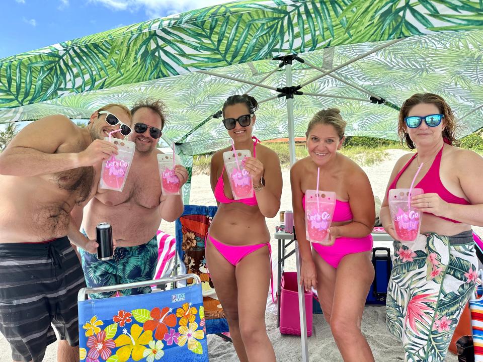 Photo of Terri Peters and her friends standing under umbrellas with a jungle leaf print. Three women on the right wear pink bikinis and hold adult Capri Suns filled with vodka and pink lemonade that have pink straws and say, "Let's Go Party." There are two men on the left who wear dark swim trunks.