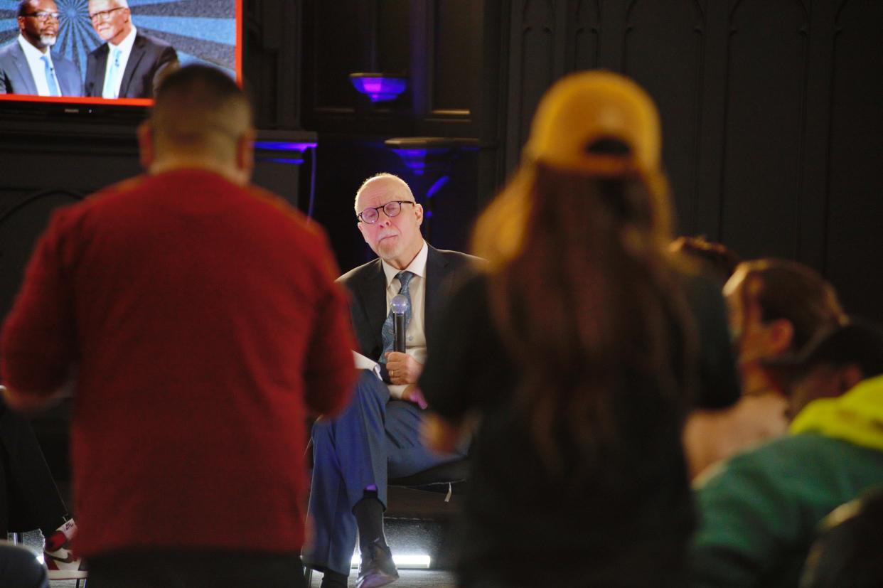 Paul Vallas stops to think onstage, surrounded by an audience, with a screen in the background showing images of him and Brandon Johnson.