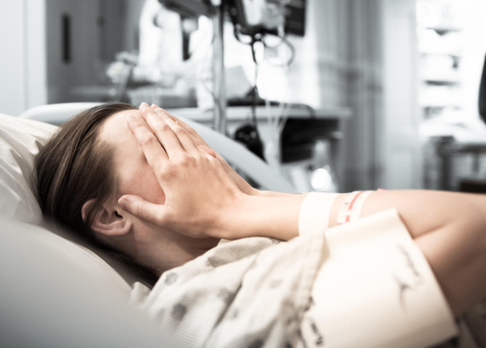 A young woman in a hospital bed with her hands covering her face. She might be in pain following a procedure like getting an IUD inserted. (Photo via Getty Images)