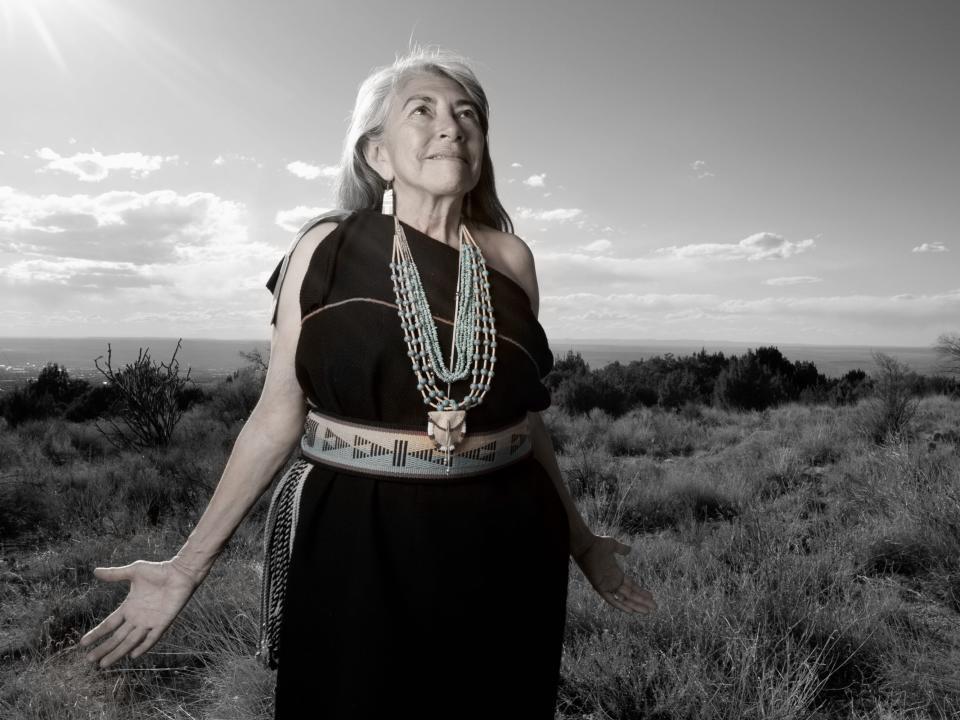 A Native woman photographed by Matika Wilbur. She looks up at the sky with her hands extended at her sides.